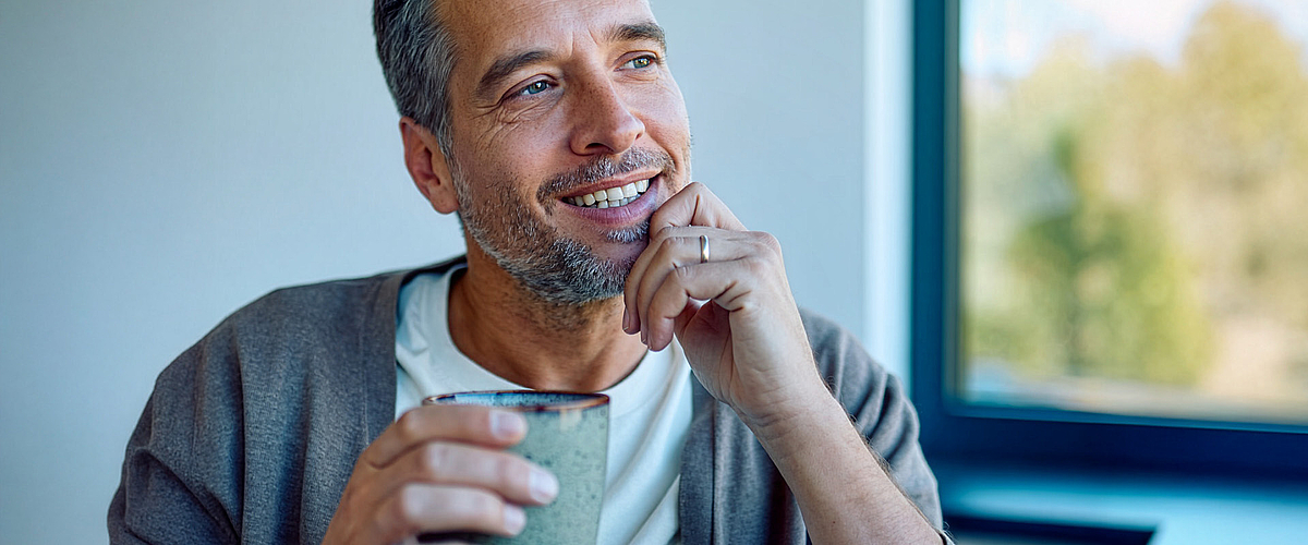 Ein Mann sitzt grüblerisch lächelnd mit einer Tasse Tee am Fenster